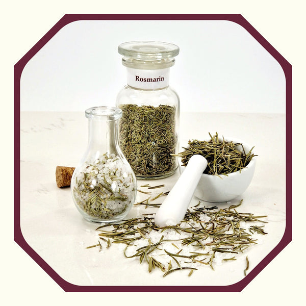 Rosmarin im Glas neben einem Glas mit Rosmarin-Peeling-Salz, einem kleinen weißen Mörser mit Rosmarinnadeln und Rosmarin und Salz