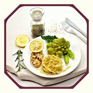 Rosmarin im Glas neben einem Teller mit einem Nudelnet, Romanesco, Rosmarin-Zitronen-Hähnchen und einer Scheibe Zitrone, einem Rosmarinzweig und Besteck