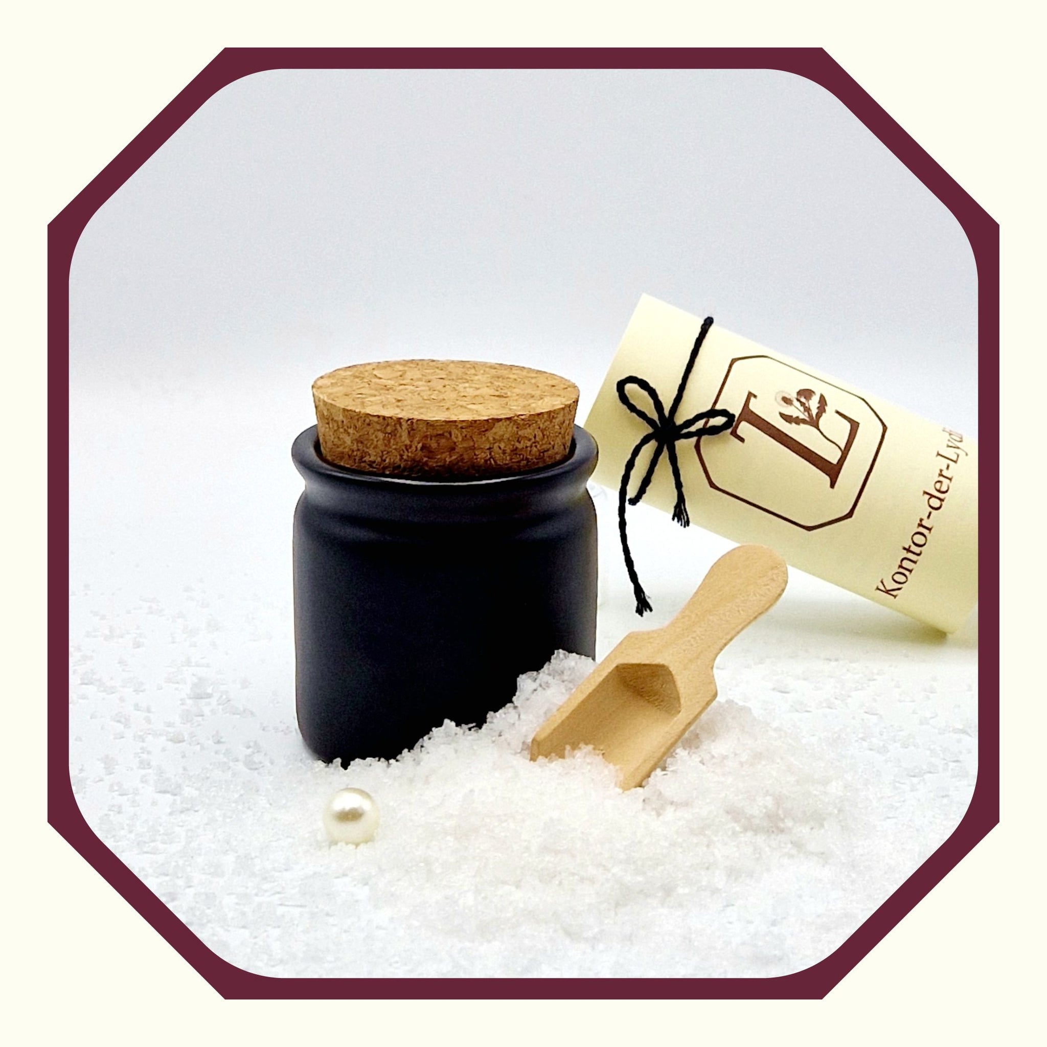 Eine Perle, eine rund gefaltete Karte mit schwarzem Schleifenband, einem schwarzen Keramik Salztopf, davor loses Fleur-de-Sel, eine kleine Salz-Schaufel aus Holz