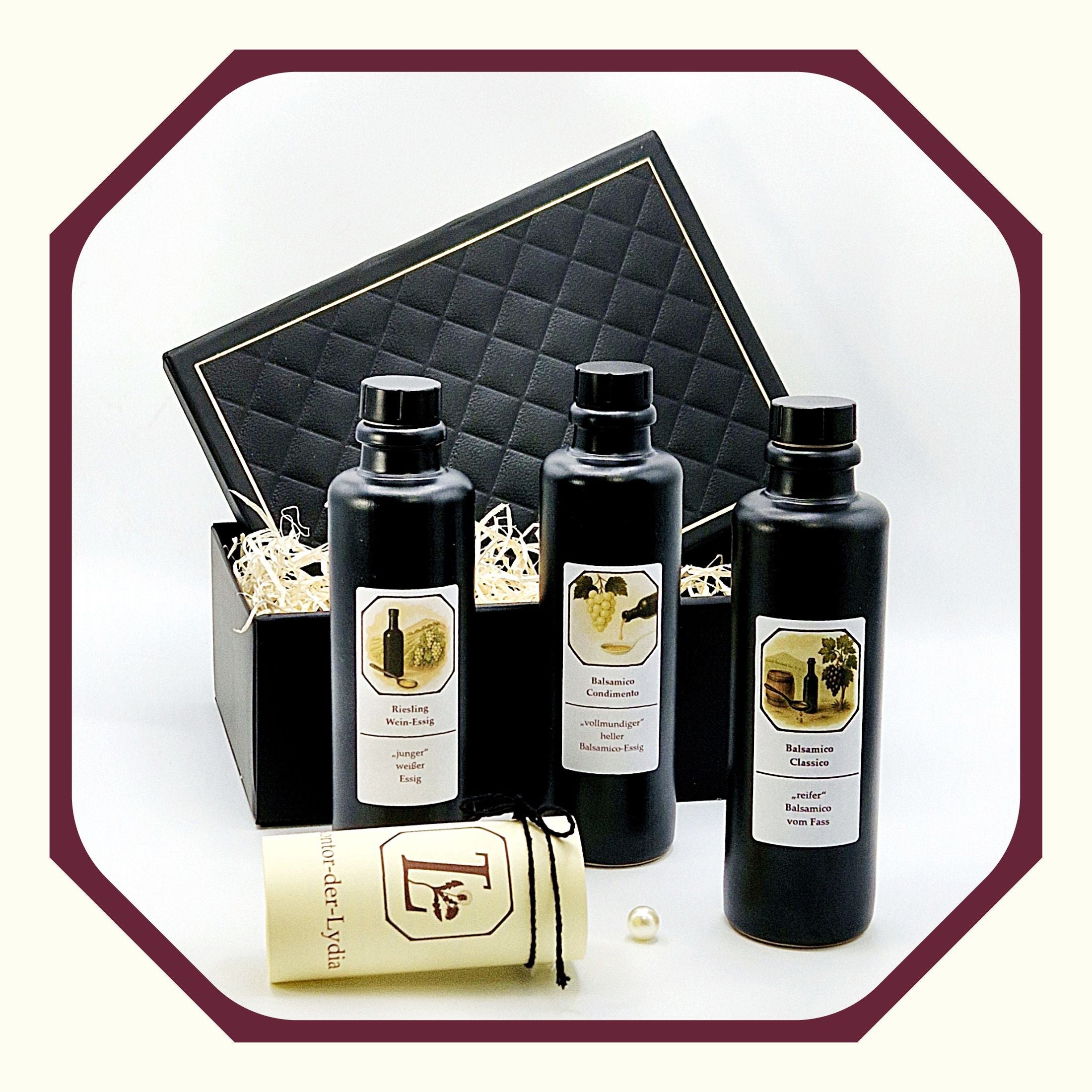Eine Perle, eine rund gefaltete Karte mit schwarzem Schleifenband, eine schwarze Tonflasche mit Essig Riesling Weinessig, eine schwarze Tonflasche mit Essig Balsamico Condimento, eine schwarze Tonflasche mit Essig Balsamico Classico vor einer schwarzen Luxus Geschenkbos
