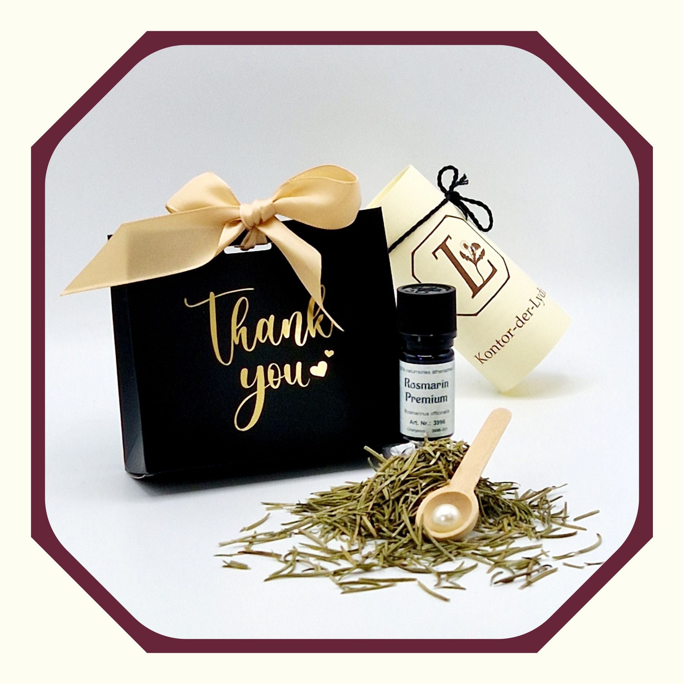 Eine Perle, eine rund gefaltete Karte mit schwarzem Schleifenband, ein Ätherisches Öl Rosmarin Premium, Rosmarin, ein Kräuterlöffel aus Holz, im Hintergrund eine kleine schwarz-goldene Geschenktasche mit dem Aufdruck Thank you und Schleife