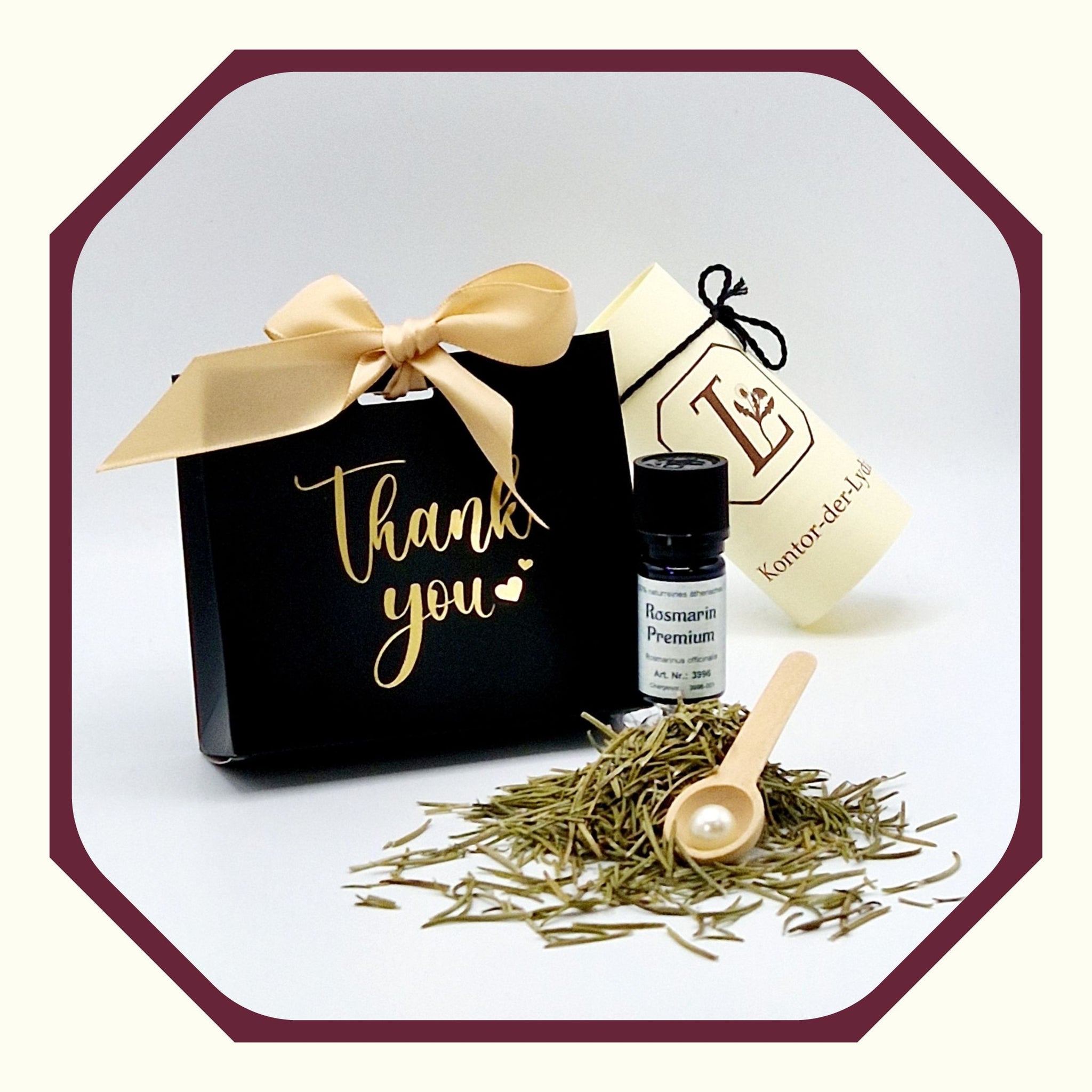 Eine Perle, eine rund gefaltete Karte mit schwarzem Schleifenband, ein Ätherisches Öl Rosmarin Premium, Rosmarin, ein Kräuterlöffel aus Holz, im Hintergrund eine kleine schwarz-goldene Geschenktasche mit dem Aufdruck Thank you und Schleife